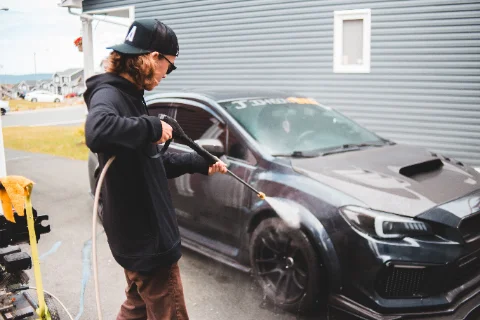 A teenager in a cap pressure-washing a black car on a driveway.