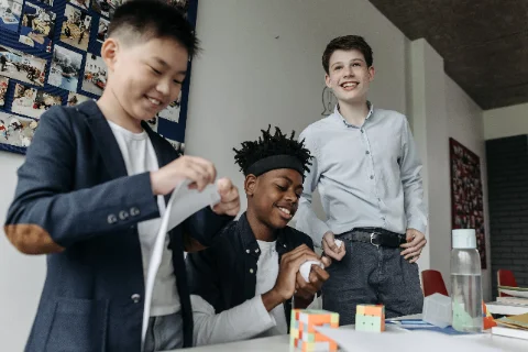 Three teenagers laughing while building something from paper and card at a table.